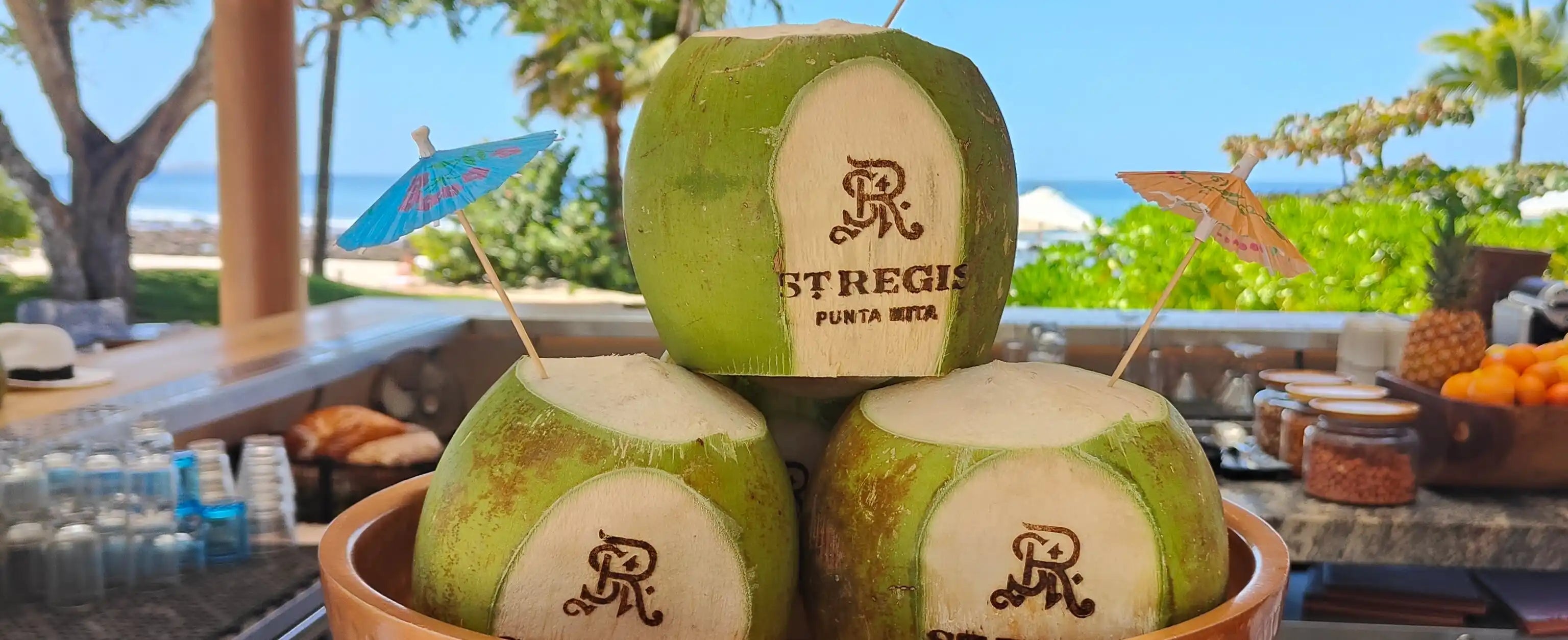 Three coconuts with the St. Regis logo branded with our custom branding irons sitting in a bowl on a table outdoors with a scenic background.