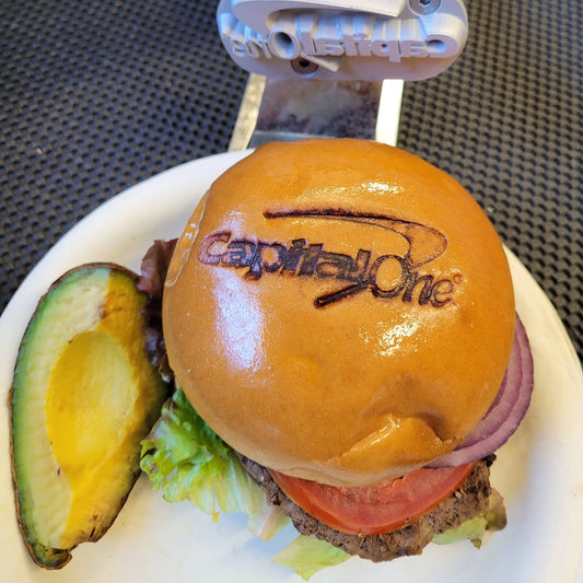 Hamburger with Capital One branding iron marked on a bun, accompanied by an avocado and tomato, on a white plate.